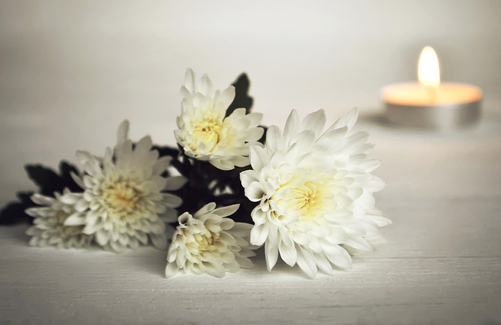 A cluster of white flowers rests on a light wooden surface, with a lit tealight candle in a metallic holder glowing softly in the background.