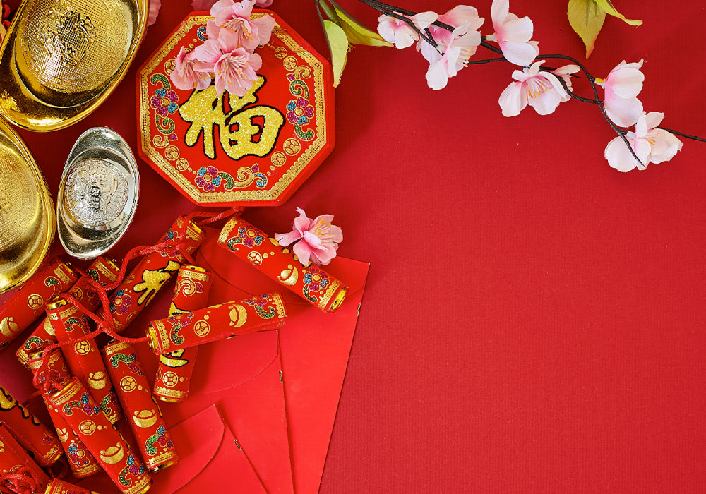 Golden ingots, firecrackers, and a red octagonal items marked with a gold Chinese character, placed on a red surface, with added decorative pink flowers.