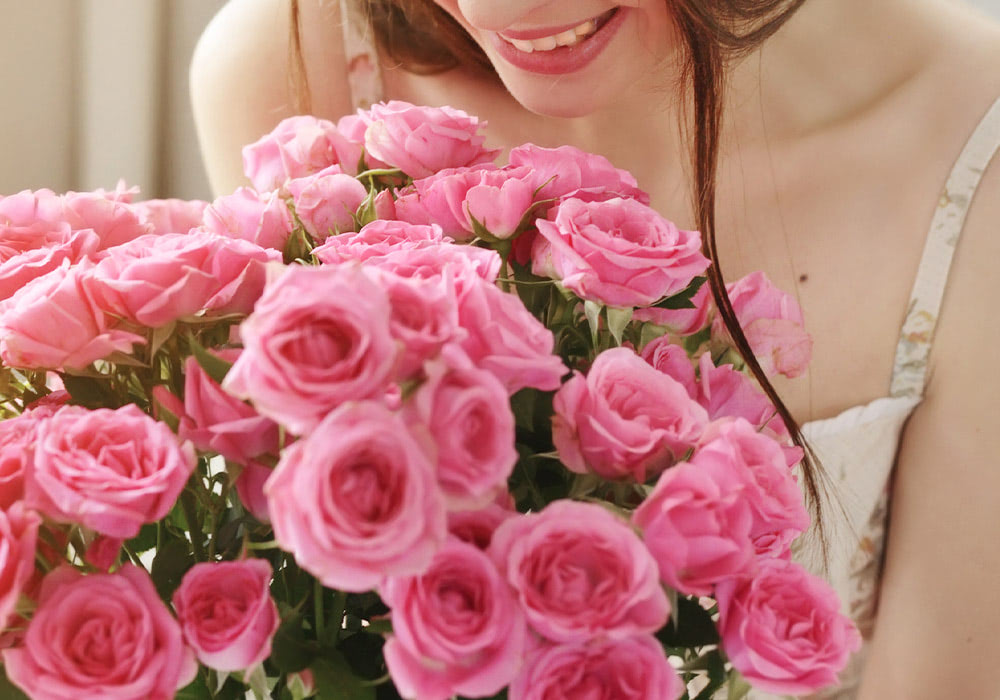 A person holding a large bouquet of vibrant pink roses, smiling while leaning in closely, with soft indoor lighting in the background.