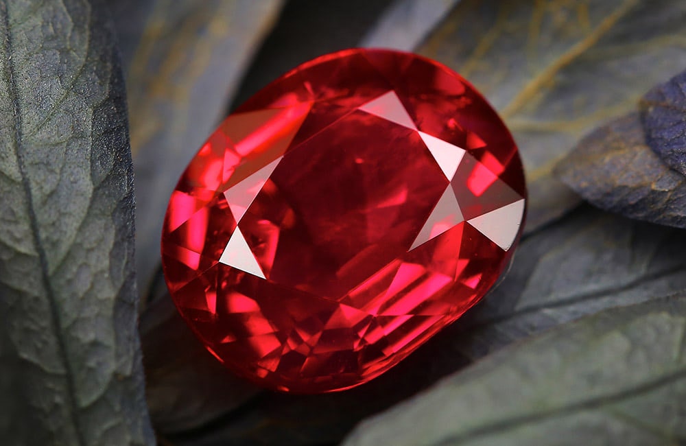 A faceted red gemstone lies on a surface of overlapping dark, matte leaves. The vibrant gem contrasts sharply with the muted background.