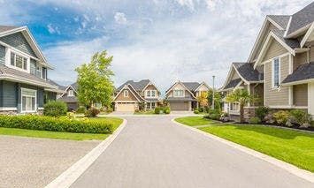 Street with well-maintained suburban houses, surrounded by green lawns and trees, under a partly cloudy sky.