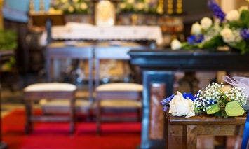 Flowers rest on a wooden pew in a church decorated for a wedding, with an altar and chairs in the blurred background. A red aisle runner leads towards the altar.