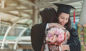 A graduate in a cap and gown hugs another person while holding a bouquet of flowers, standing under an arched metal and glass structure.