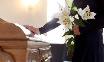 A person in black clothing holds white lilies with one hand, while the other hand rests on a wooden casket in a well-lit room.