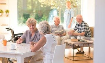 Elderly individuals are gathered in a well-lit, comfortable room with large windows. Two women converse at a table, while three men sit and chat in the background, accompanied by a caregiver.