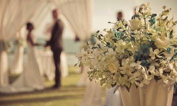 A lavish bouquet of white and blue flowers sits in the foreground while a couple stands under a white canopy exchanging vows in an outdoor wedding ceremony.